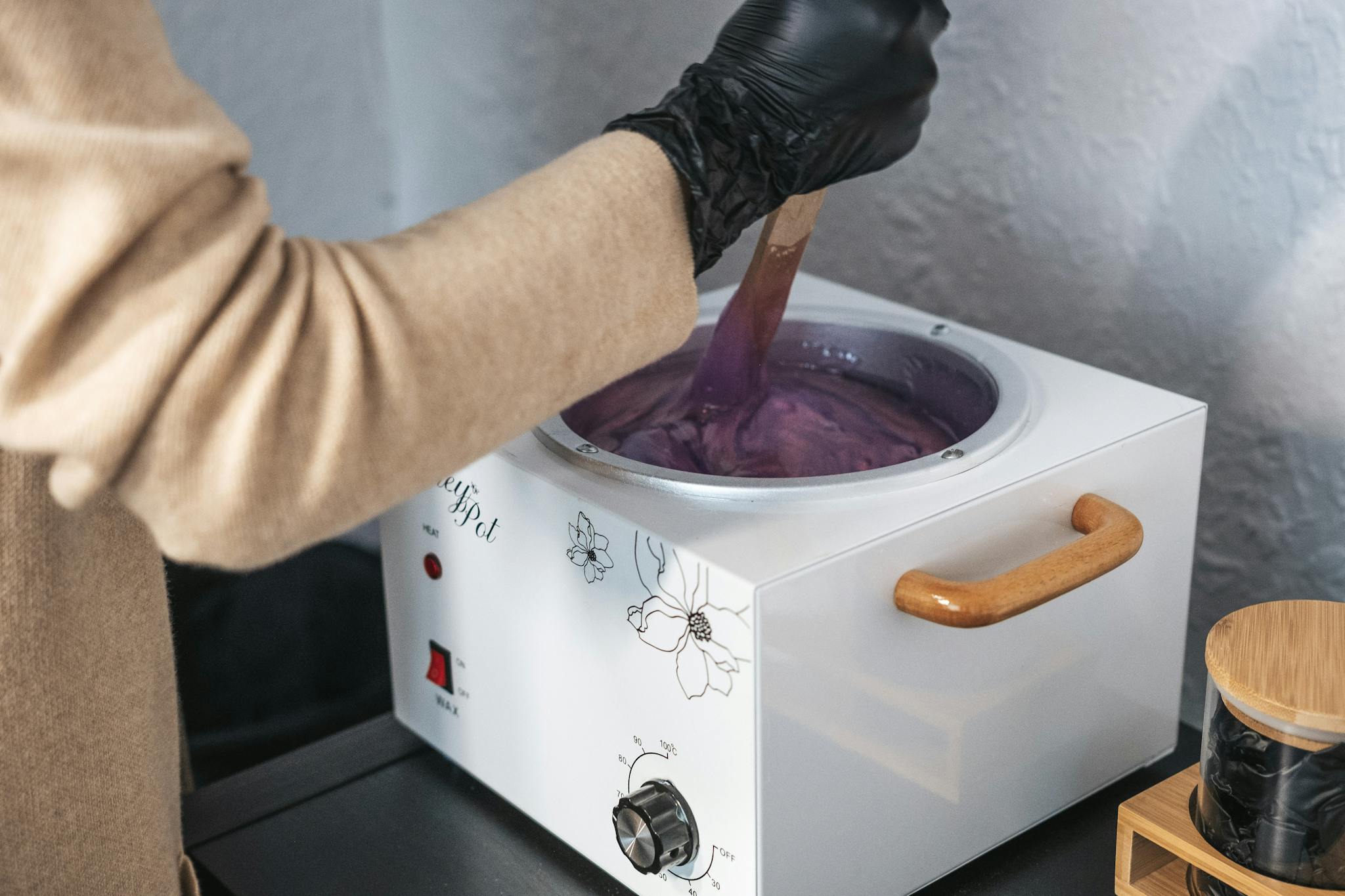 An esthetician wearing gloves stirring purple wax in a wax pot indoors.